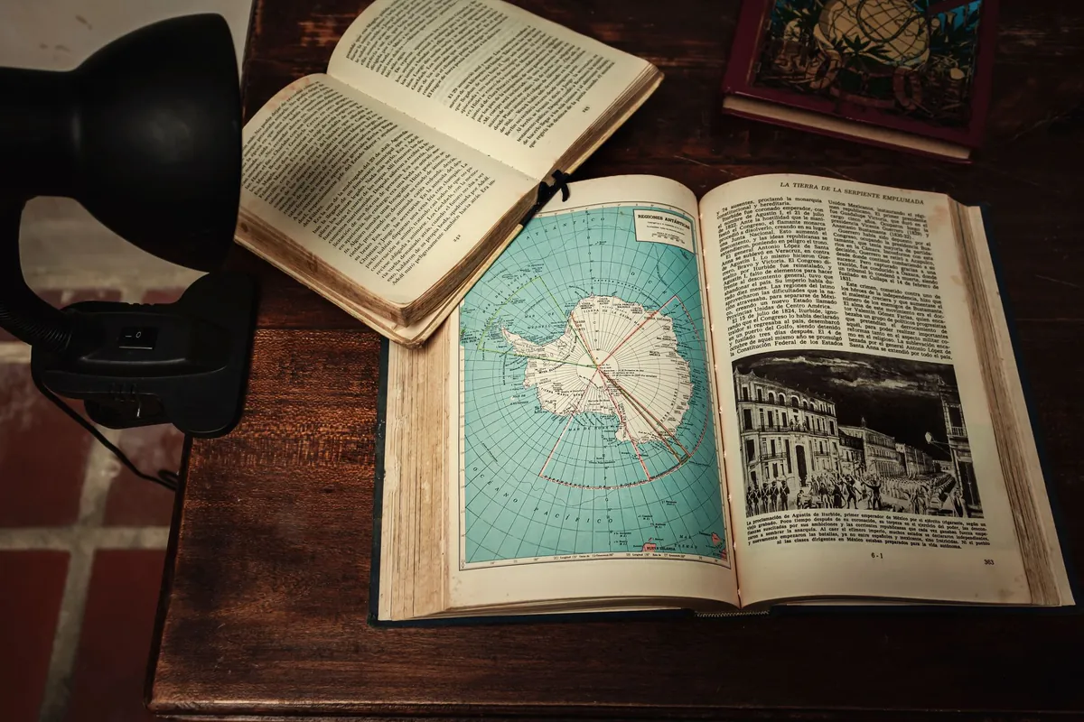 Old books open on a wooden desk under a black desk lamp, one showing a map of Antarctica